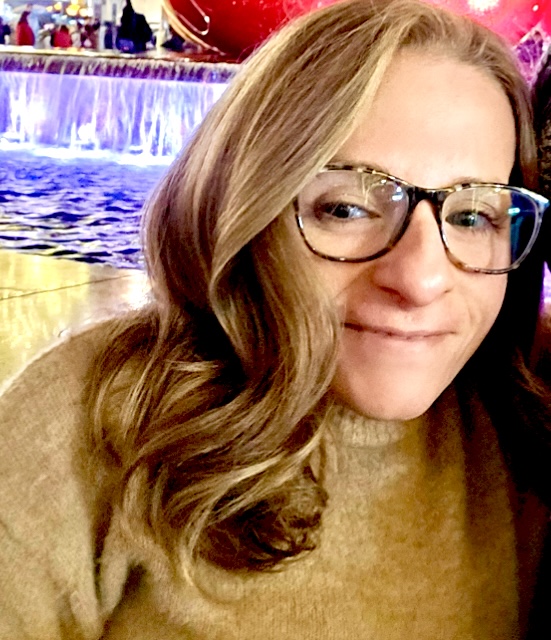 Heather is looking at the camera smiling. She is wearing a tan sweater, her hair is dark blonde and in curls, and she is wearing tortoise patterned eye glasses.